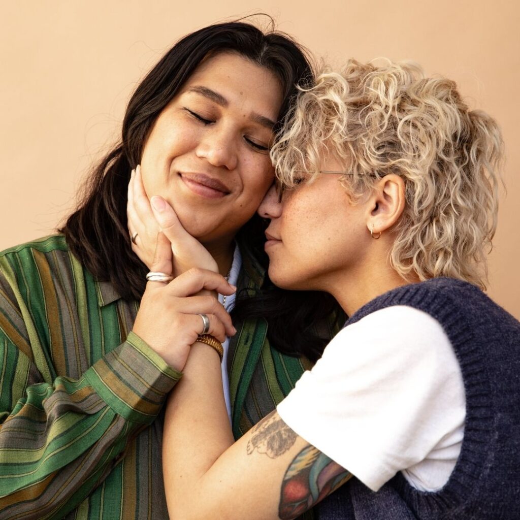 Two non-binary presenting people are cuddling outside against a tan wall. The person on the left has long brown hair and a green shirt. The person on the right has curly blonde hair and tattoos.