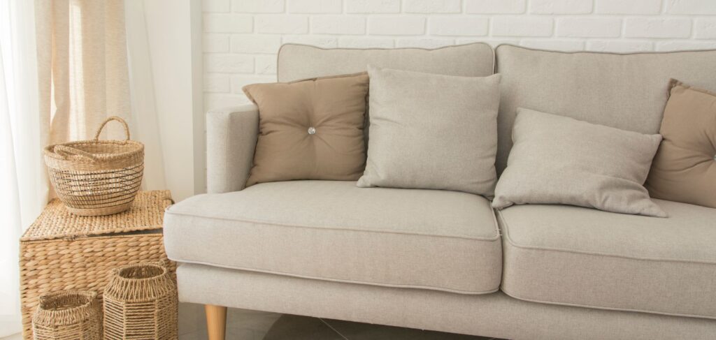 A grey therapy couch with four pillows on it and a white brick background. It is wide enough for multiple fat bodies.