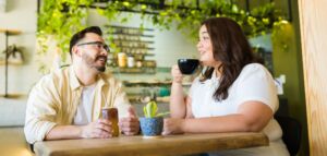 Couple in a bright restaurant sharing drinkings and smiling. The woman is in a bigger body and he looks at her adoringly.