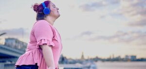 Plus size young white woman in a pink shirt wears headphones and leans into the sunshine on a seaside railing.
